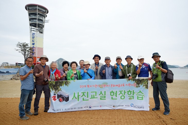 군산선유도 해변에서 어르신과 강사 총 13명이 손으로 하트모양을 만들며 웃음을 띄고 단체사진을 찍고 계신다   